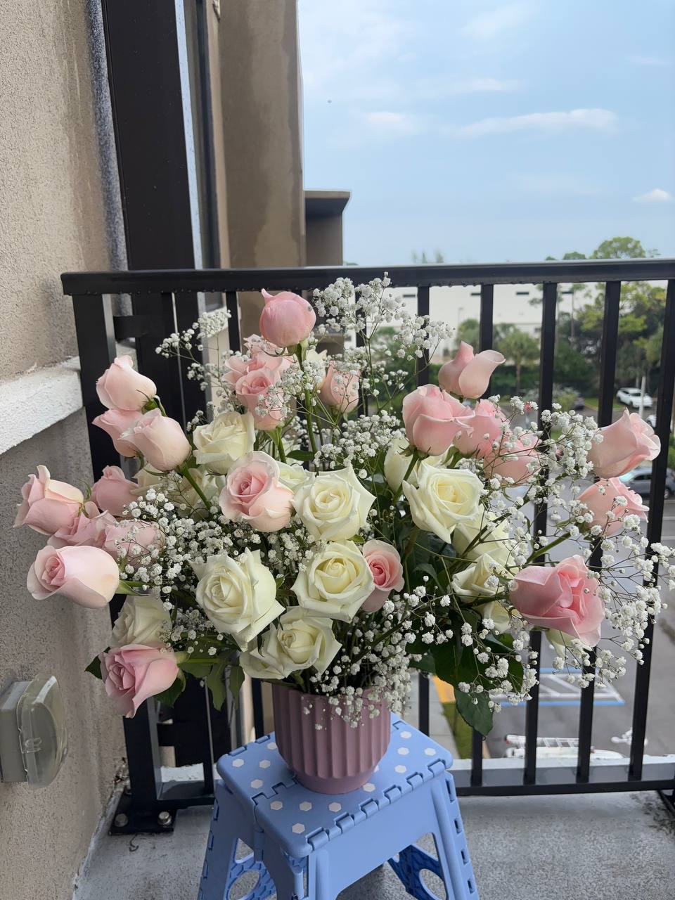 Tall arrangement of pale pink and white roses with baby's breath in a ribbed pink vase, set on a balcony