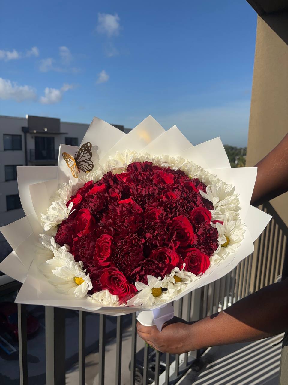 Red roses and carnations ringed with white daisies in white wrapping, with a gold butterfly accent