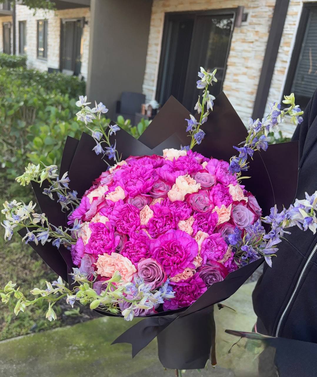 Magenta carnations, peach carnations and mauve roses with lavender delphinium in black wrapping